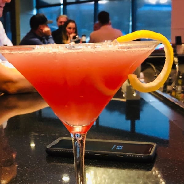 A pink cocktail with a lemon twist on a bar counter, with blurred patrons in the background.